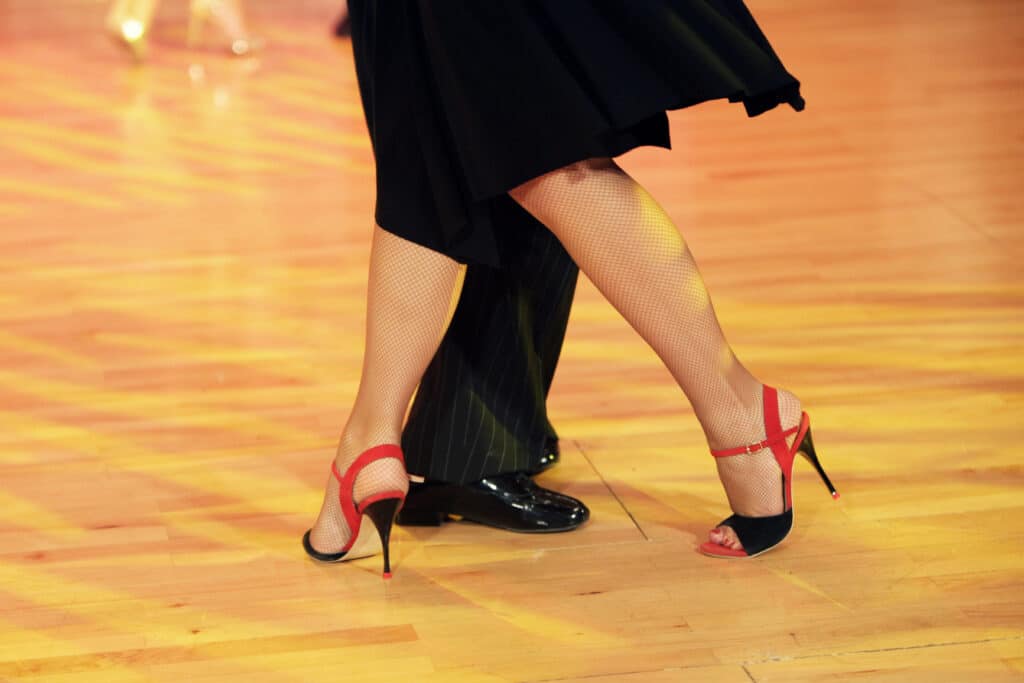 Pieds élégants de danseurs de salon: femme en talons rouges et bas résille, homme en chaussures noires sur parquet clair.