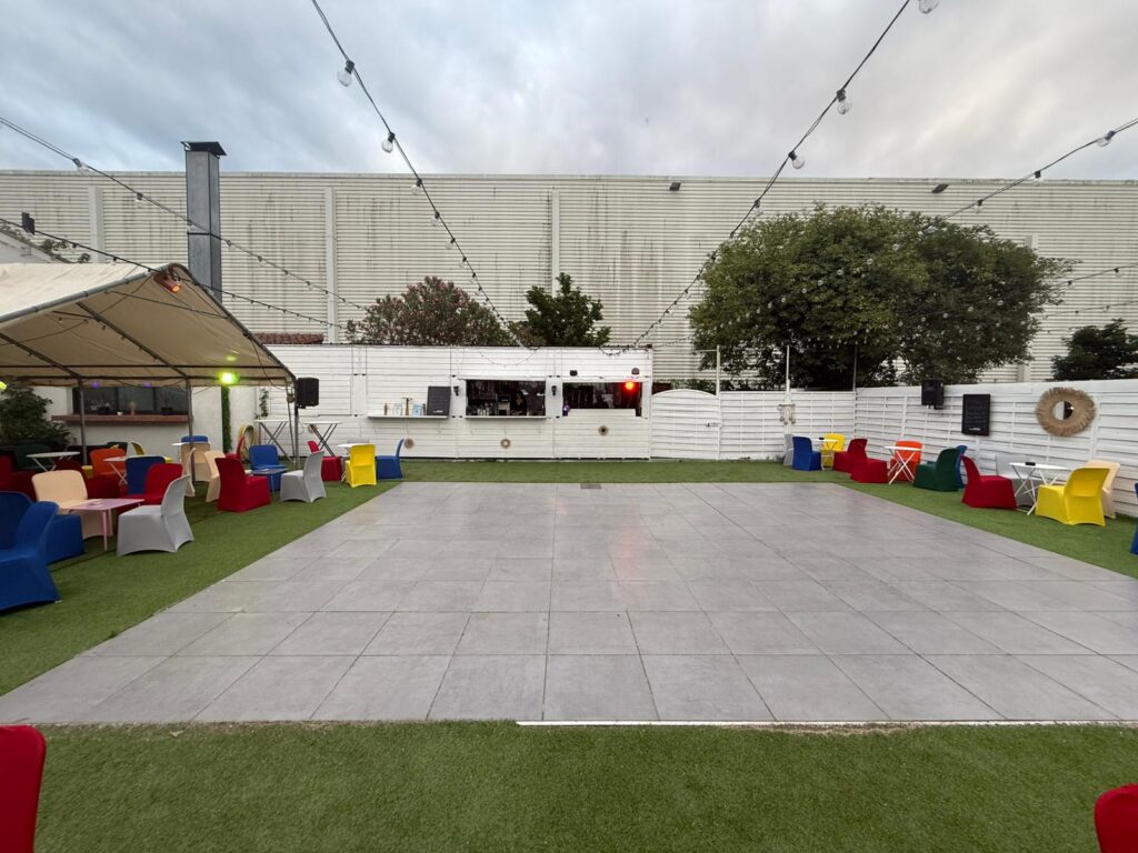 Terrasse événementielle avec piste de danse et bar Terrasse événementielle avec piste de danse grise, fauteuils colorés, bar blanc et guirlandes sous un ciel nuageux.